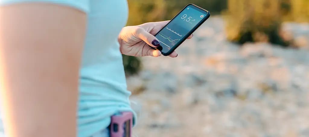 Frau hält Handy mit Blutzucker-Messgerät in der Hand. Im Hintergrund ist Gebüsch zu sehen.